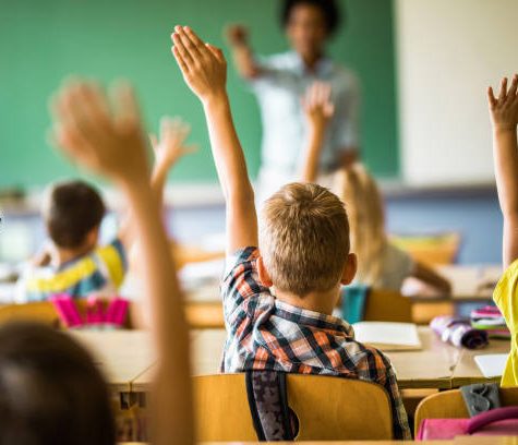 istockphoto-1223141903-612x612 Rear view of large group of students raising their hands to answer the question on a class at elementary school.