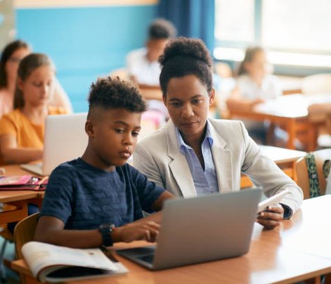 istockphoto-1331594985-612x612 Black female teacher assisting schoolboy in using laptop during a class in the classroom.